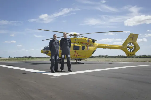 Captain James Pusey (on left) and Pilot Prince William (on right) in flying uniform standing in front of air ambulance