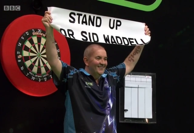 Phil Taylor holds a sign saying "stand up for Sid Waddell"