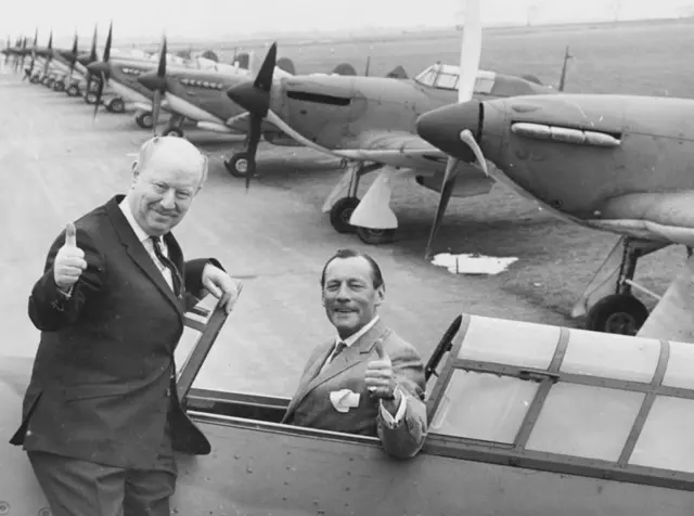 Former fighter pilot Wing Commander Robert Stanford-Tuck sitting in the cockpit of Hurricane aircraft giving the thumbs up, with fellow flying ace James Harry Lacey,