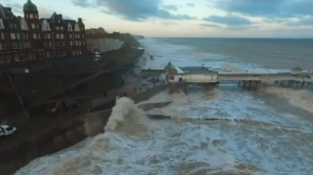 Aerial view of the promenade, showing waves crashing, and the pier in background