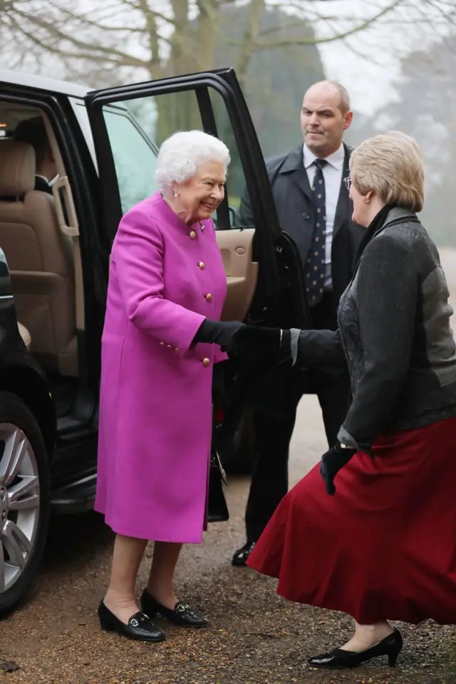 The Queen, in bright cerise coat and black shoes extending her hand to Yvonne Brown, who is curtseying