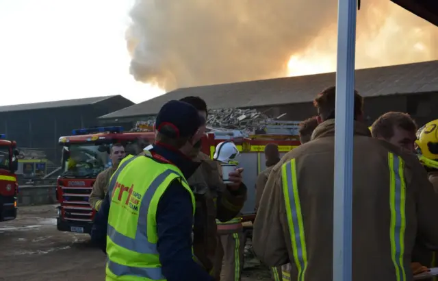 Refreshments on Sunday, firecrews standing near fire engines, with smoke billowing in background