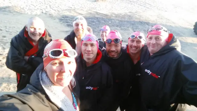 Swimmers, with goggles and wearing coats, stand on Sea Palling beach