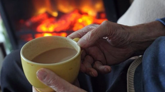 Elderly woman sitting beside fire with cup of tea