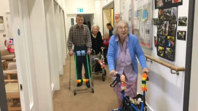 Three residents uses their frames as they walk along a corridor