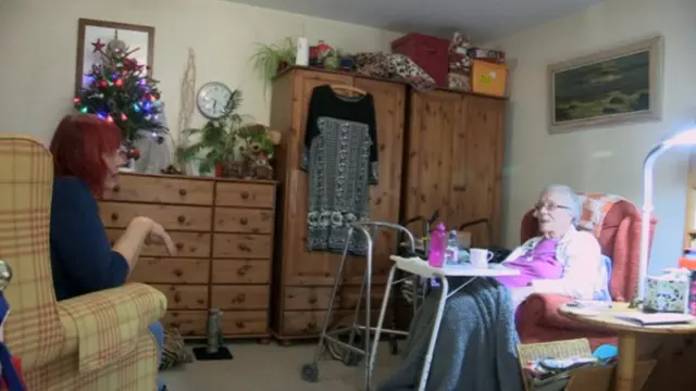 Sue sitting opposite her mother, who's in an armchair with a blanket over her legs, and a walking frame nearby