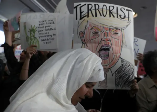 A woman wearing a headscarf walks in front of a sign borne aloft, which features a cartoon rendition of Mr Trump and the word "terrorist"