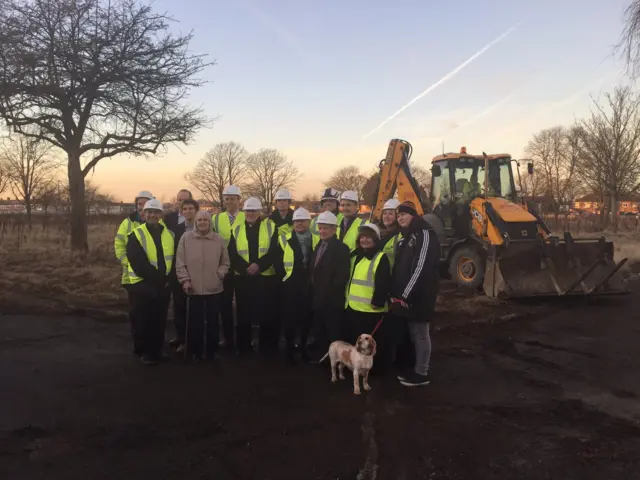 Group of officials in high-vis and hard hats standing in front of a digger