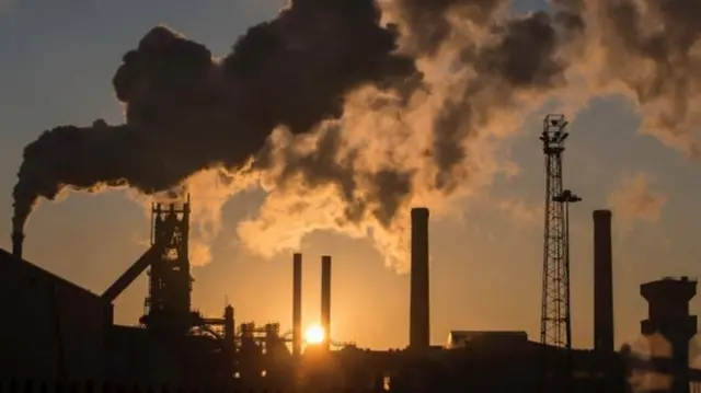 Chimneys at the steelworks with smoke coming out of then and the sun setting behind them.