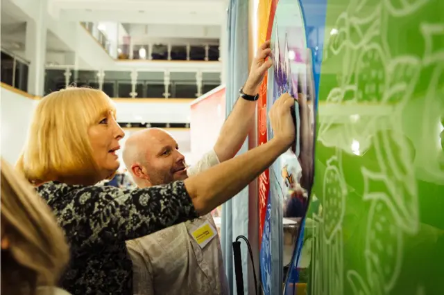 People touching a large colourful polyresin picture with the outlines and textures of animals and plants on it.