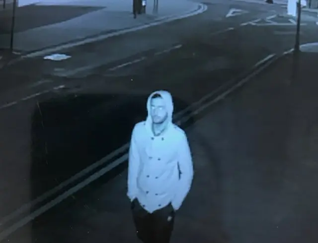 CCTV image of a man in a light coat with a hood and dark trousers