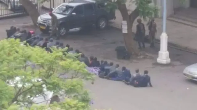 A line of police officers sit on the ground under the supervision of nearby soldiers