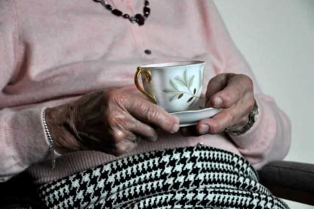 An elderly woman sits, holding a china cup and saucer