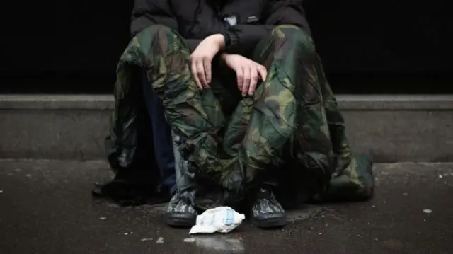 A man with a sleeping bag sits on a rainy street