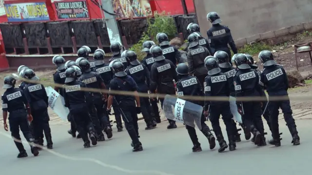 Two dozen police in body armour, helmets, and shields walk down a street away from the camera