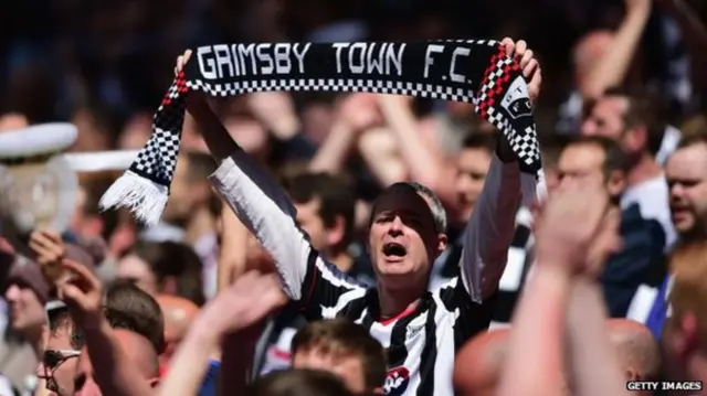 Man holding Grimsby Town FC scarf in the air at a match.