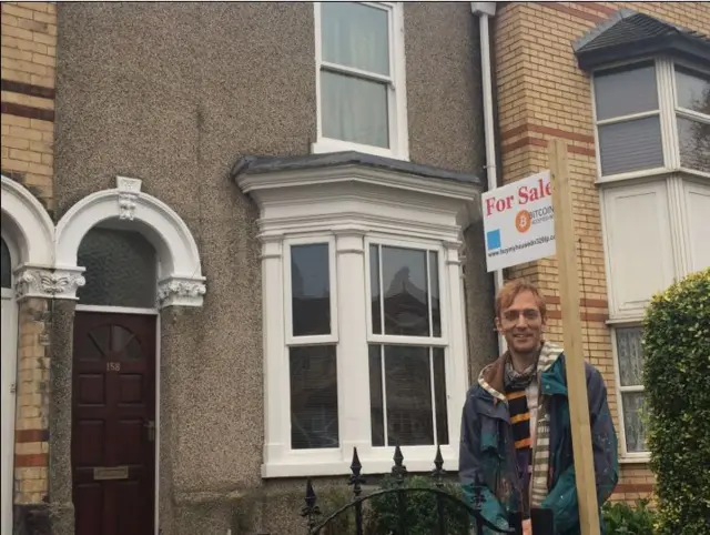 Sean outside his house with a sign saying for sale, 8 bitcoins.