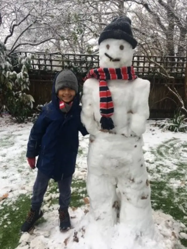 A child standing with a large snowman he built with his family and neighbours