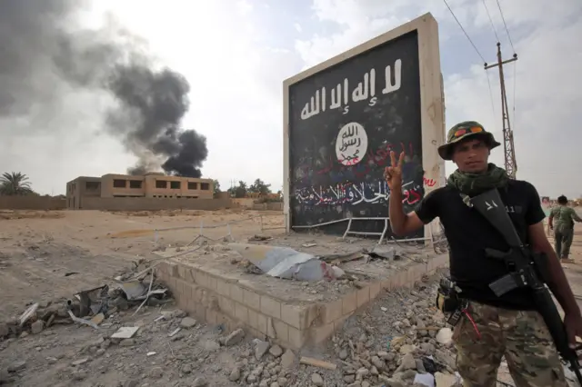 Iraqi fighters of the Hashed al-Shaabi (Popular Mobilisation units) stand next to a wall bearing the Islamic State (IS) group flag as they enter the city of al-Qaim, in Iraq's western Anbar province near the Syrian border as they fight against remnant pockets of Islamic State group jihadists on November 3, 2017. /