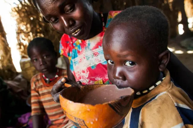 Nybol Madut (C) who lost three children to a militia, offers a bowl of local alcohol to her two-year-old son Onang Ngor (R) to satisfy his hunger, as they rest in a shelter at an Internally Displaced Person (IDP) camp