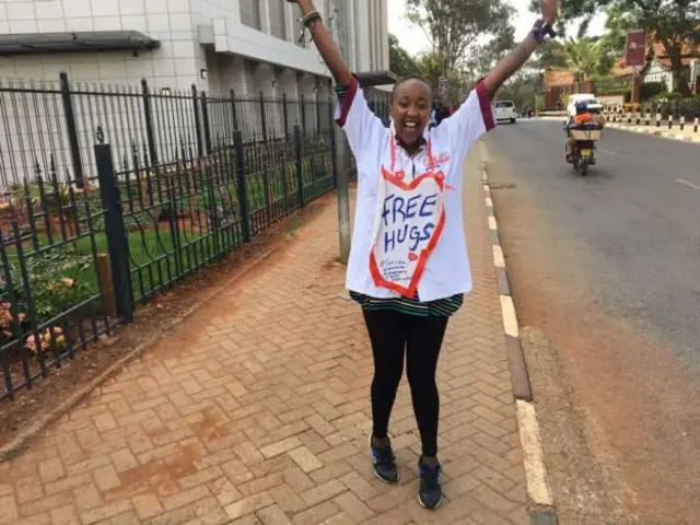 Njeri Muthaka stands on the pavement wearing a t-shirt with the message "free hugs" written inside a heart