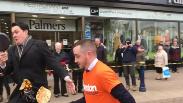 
          The two runners nearing the finishing line, tossing their pancakes, running to left of photo
        