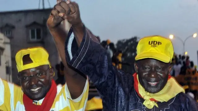 
          Ousainou Darboe (R) pictured with hand raised wearing yellow UDP baseball cap
        