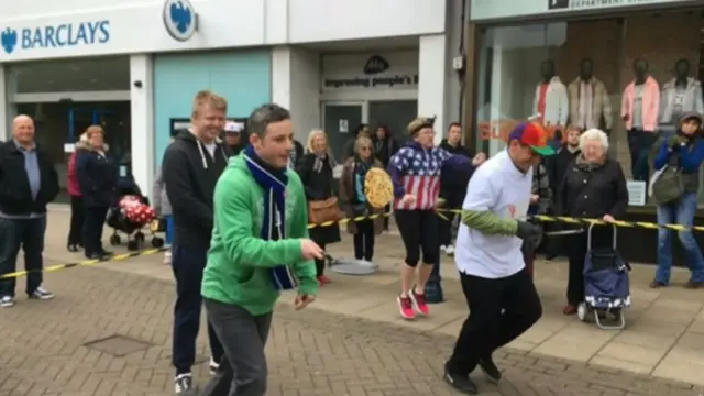 
          Two men, one in white top and multi coloured cap, the other in green top and blue scarf, set off on pancake race running to right of photo
        