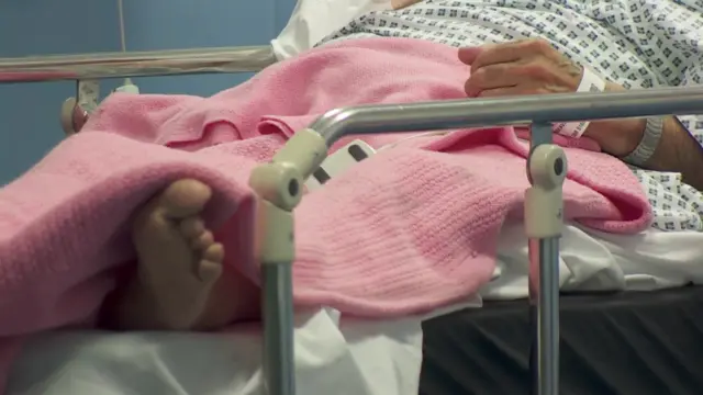 A patient in a hospital bed, wearing hospital gown with a pink blanket over their legs