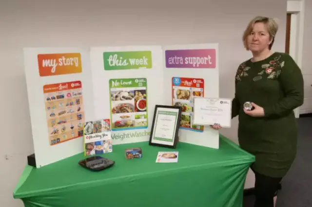 Sandra Forder holding certificate, in front of advertisements for Weight Watchers