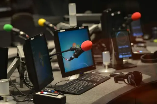 A radio studio with a red microphone in the foreground and computer screens in the background