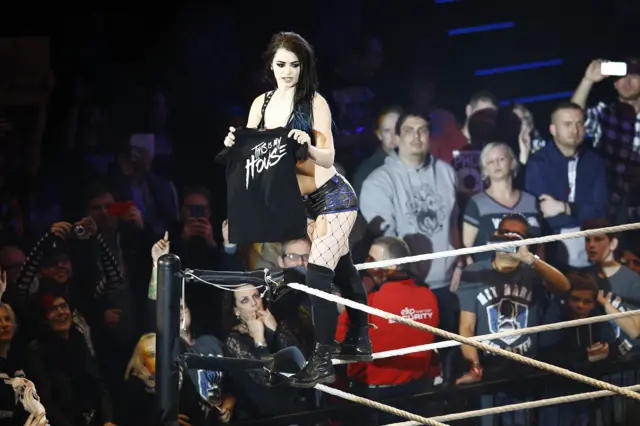 Paige in wrestling ring, in black top and shorts, holding a black tee-shirt