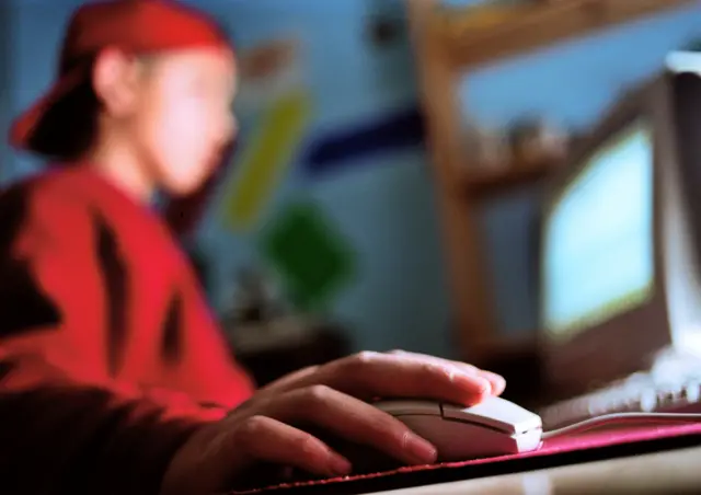 
          Blurred image of a boy wearing red top and red cap, sitting at a computer at home
        