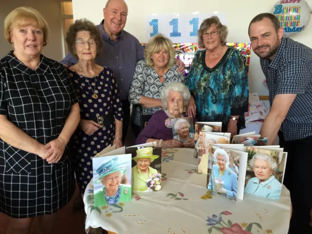 
          Family and friends standing, with Francis Malin seated, around a table with birthday cards featuring the Queen
        