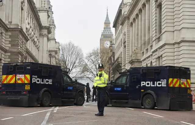 
          Police officer in front of parked armed police personnel vehicles
        