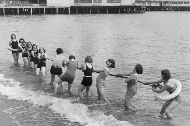 April 1933: A group of girls playing tug-o-war in the sea next to the pier at Clacton, Essex.