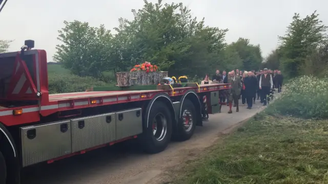 Coffin with orange flowers on the back of HGV, with funeral procession behind