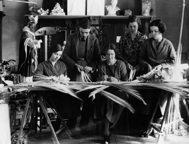 
          Black and white photo showing workers seated or standing around a table, making palm crosses
        