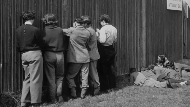 Children watch horseracing through gaps in the fence