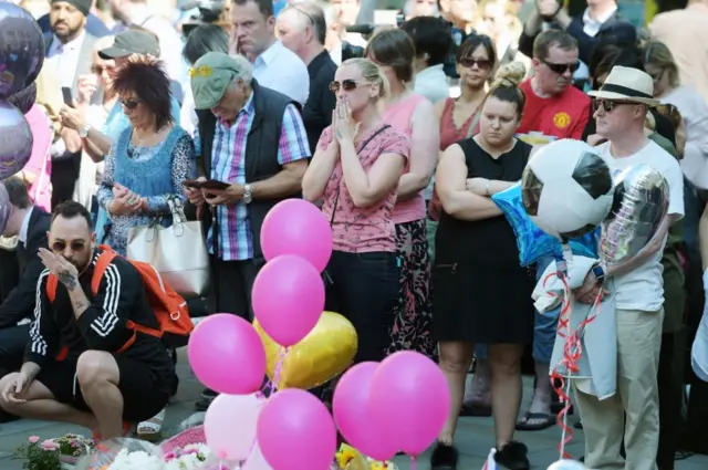 People stand by floral tributes in St Ann's Square, Manchester, ahead of a minute's silence