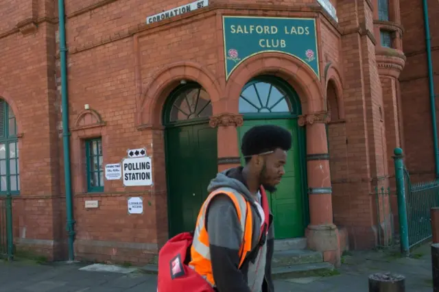 A postman walks past a polling station sign on the Salford Lads Club on Coronation Street where voting in the Greater Manchester mayoral election is taking place,