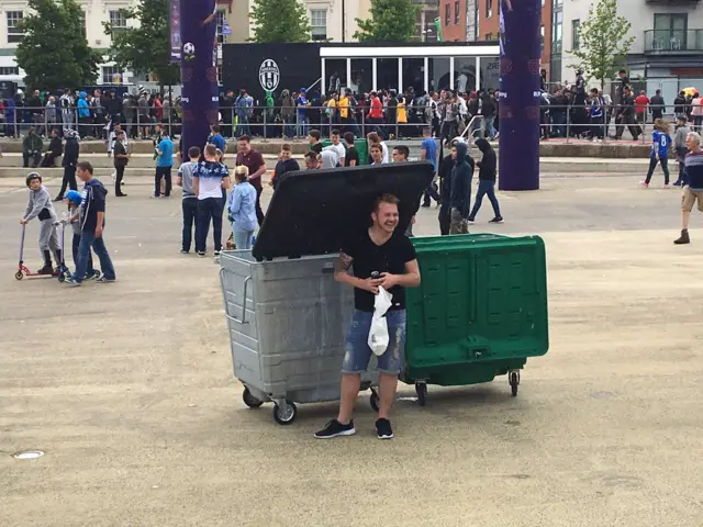 A fan takes shelter under a wheelie bin