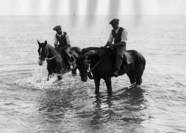 Black and white photo of two horses, ridden by stable lads, in the sea