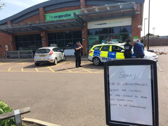 Police officers and vehicles outside supermarket plus sign saying store is closed