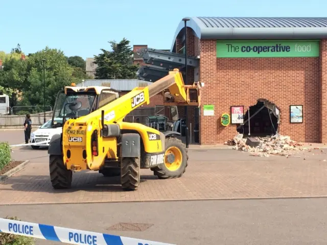 Police tape around yellow JCB teleporter, and hole in the wall of supermarket