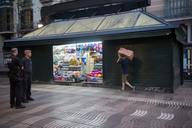 Police officers look on as a man carries a box of goods to his shop on Las Ramblas