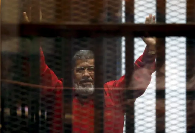 Deposed President Mohamed Mursi greets his lawyers and people from behind bars at a court wearing the red uniform of a prisoner sentenced to death, during his court appearance with Muslim Brotherhood members on the outskirts of Cairo, Egypt, June 21, 2015.