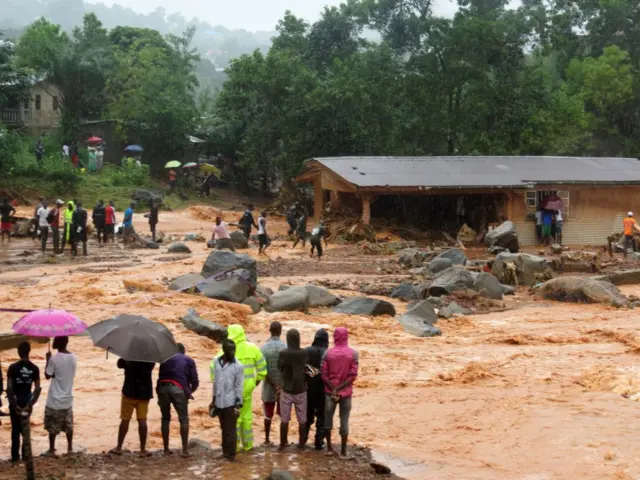 Bystanders look on as floodwaters rage past a damaged building in an area of Freetown on August 14, 2017, after landslides struck the capital of the west African state of Sierra Leone.