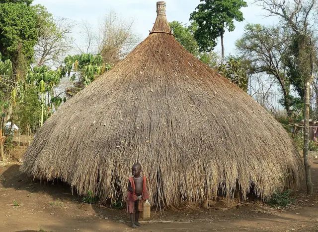 A boy stands in front of a thatched hut