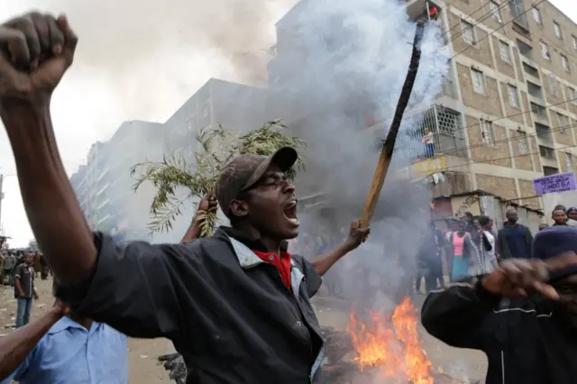 A supporter (L) of the opposition leader Raila Odinga who leads The National Super Alliance (NASA) coalition, holds a burning stick next to a burning barricade during a protest after Odinga announced that he rejects the provisional result of the presidential election announced by the electoral body, in Mathare slums one of Odinga"s strongholds in Nairobi,